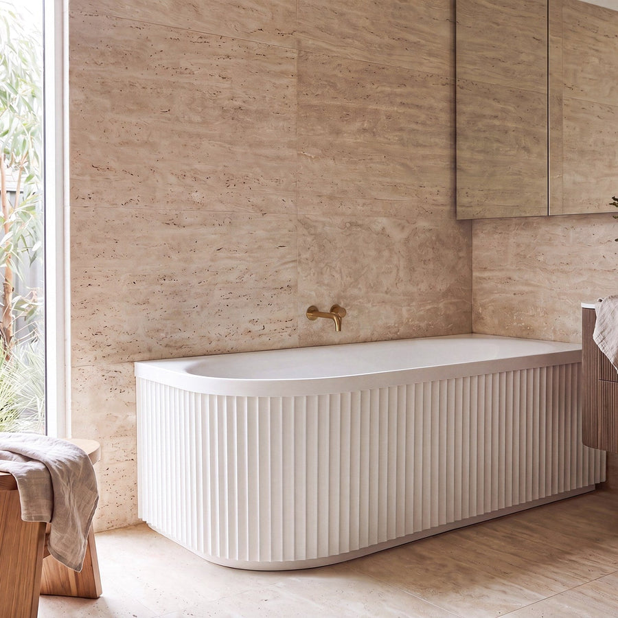 Fluted white corner bath with brushed brass spout and travertine look wall tiles