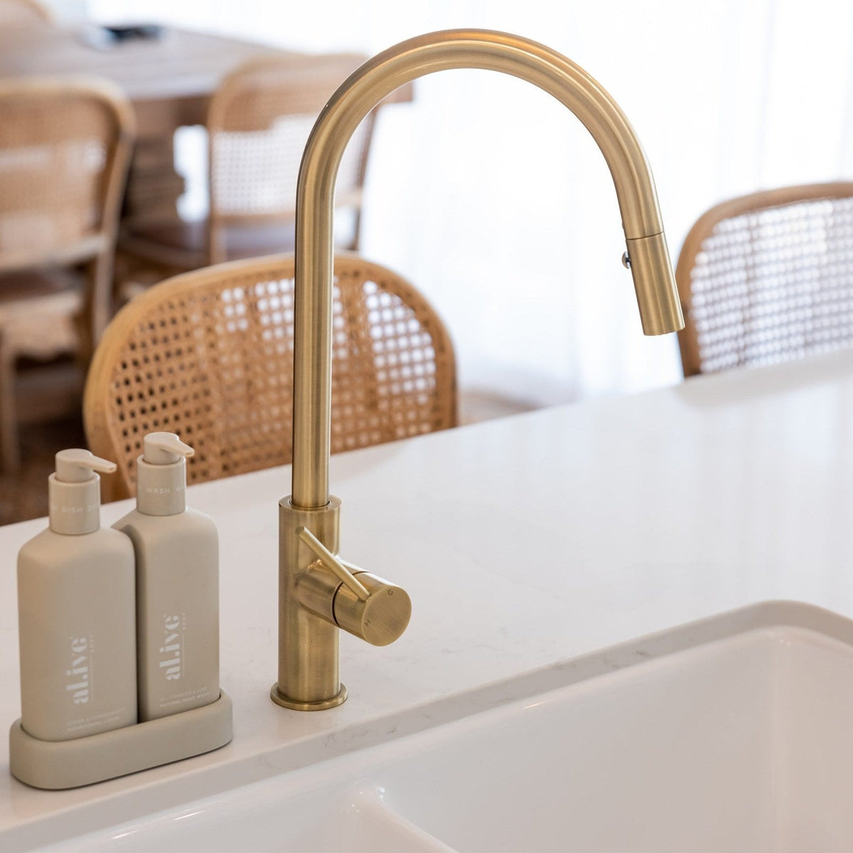 Brushed brass gooseneck kitchen mixer on a white stone benchtop with timber chairs behind