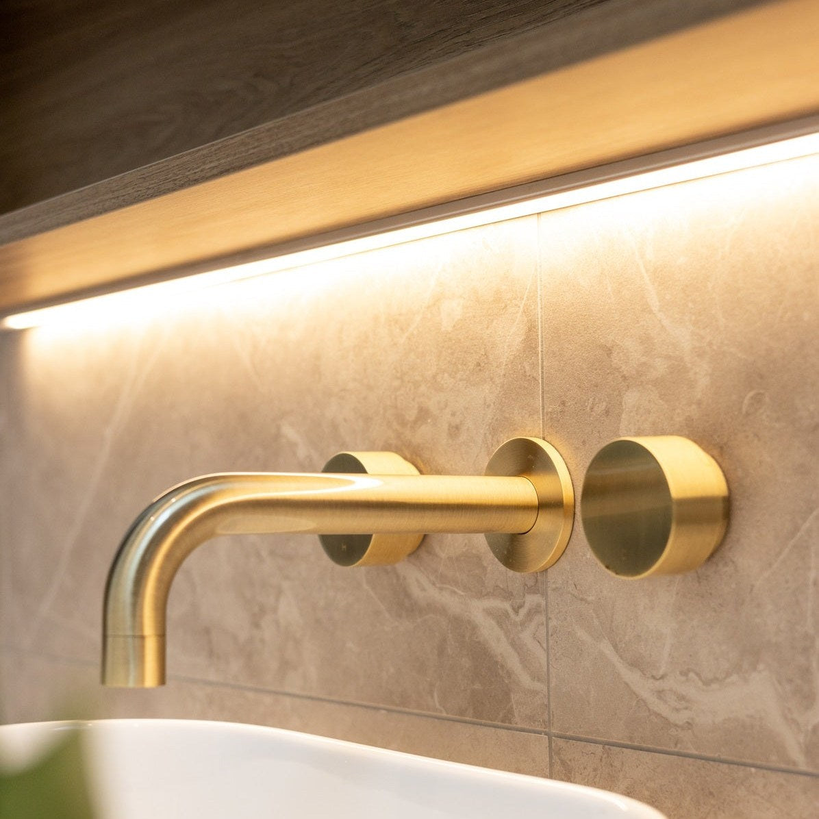 Brushed bronze wall basin taps above a stone basin in a warm, softly lit bathroom